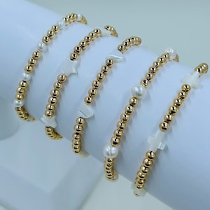 May include: Four gold beaded bracelets with white shell charms. The charms include a star, a heart, a cross, and a teardrop shape.