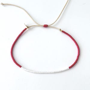 May include: A red and white beaded bracelet with a sliding knot closure. The bracelet features a section of red beads and a section of white beads. The adjustable cord is a light beige color.