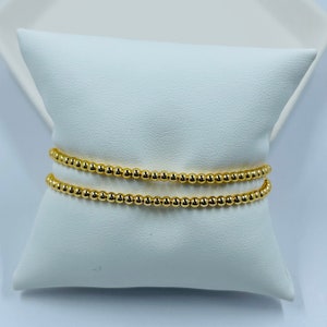 May include: Three gold beaded bracelets stacked on a white pillow. The bracelets are made of small, round beads.