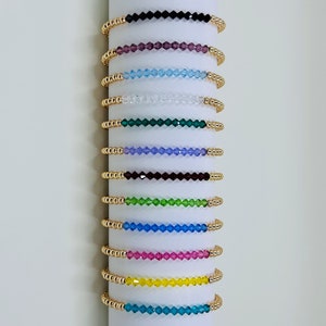 May include: A set of 12 gold beaded bracelets with different colored crystal beads. The bracelets are stacked on a white background.