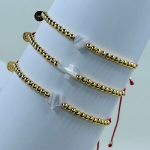 May include: Three gold beaded bracelets with red string closures. Each bracelet features a white charm, including a cross, a heart, and a wave.