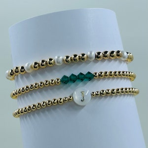 May include: Four gold bead bracelets stacked on a white surface. The top bracelet has white pearls and green crystals. The bottom bracelet has a white pearl charm with the letter "J" on it.