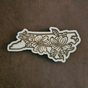 Puede incluir: Un recorte de madera del estado de Carolina del Norte con un dise&ntilde;o floral. Las flores son estilizadas y se asemejan a flores de cornejo.