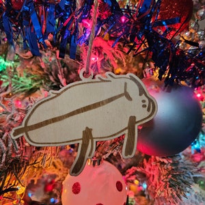 May include: A wooden Christmas ornament shaped like a pig, hanging on a Christmas tree with red, blue, and white tinsel. The ornament is painted in a light brown color and has a simple, cartoon-like design.