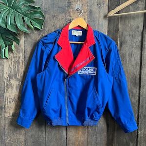 Puede incluir: Una chaqueta azul y roja con cierre de cremallera. La chaqueta tiene un cuello rojo y las palabras "SKYLINE Racing Team" están bordadas en la parte delantera.