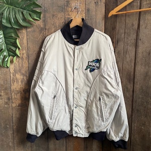 May include: A light grey bomber jacket with black ribbed cuffs and collar. The jacket has a zipper closure and a button on the front. It features a large embroidered patch on the left chest that says "HKS" in white letters with a blue and green graphic design.