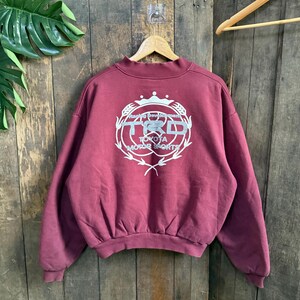 May include: A maroon sweatshirt with a crew neck and long sleeves. The back features a white graphic of the Toyota Motor Sports logo, including the letters "TRD" within a laurel wreath design. The sweatshirt is hanging on a wooden hanger.