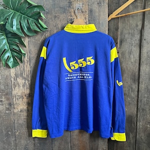 Puede incluir: Una camisa azul de manga larga con detalles amarillos. La camisa tiene un cuello amarillo y rayas amarillas en las mangas. La parte posterior de la camisa tiene un gran gráfico amarillo con el texto "555 Smoothness Above All Else."