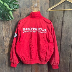 May include: A bright red long-sleeve jacket featuring a high collar and elasticized cuffs and hem. The back displays the white text Honda Racing Team, and a small white Honda logo patch is visible on the right sleeve near the shoulder area.