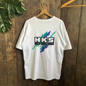 May include: White t-shirt with a graphic print of the word "HKS" in black and white with a green, blue, and red abstract design behind it.