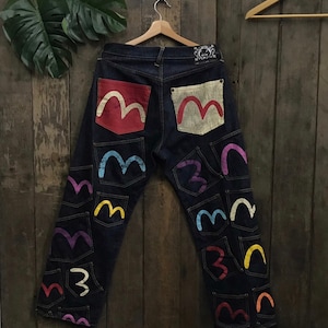 Puede incluir: Vaqueros de mezclilla azul oscuro con múltiples bolsillos bordados de colores en forma de una 'M' estilizada.