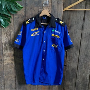 Può includere: Una camicia blu a maniche corte con bottoni, colletto e polsini neri. La camicia presenta diversi loghi, tra cui "Subaru", "Motul" e "555" in lettere gialle e dorate.