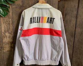 May include: A gray and white windbreaker jacket with a red stripe and the words "RALLI ART" printed on the back. The jacket has a Mitsubishi Motors logo on the left sleeve.