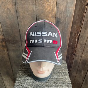 May include: Black baseball cap with red and white accents. The cap has the words "NISSAN nismo" embroidered on the front.