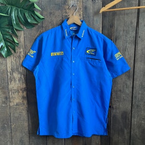 Può includere: Una camicia blu a maniche corte con bottoni e loghi ricamati in giallo, tra cui "Subaru", "Pirelli" e "STI".