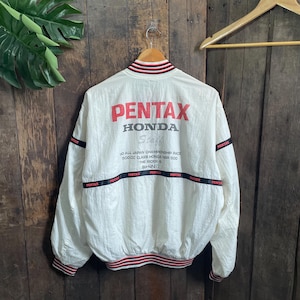 May include: A white long-sleeve jacket with red and black striped ribbed trim at the collar, cuffs, and hem. The back features red and black text reading PENTAX HONDA Staff, 90 ALL JAPAN CHAMPIONSHIP RACE, 500CC CLASS HONDA NSR 500, THE RIDER IS SHINYA.