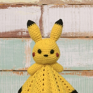 May include: A yellow crocheted Pikachu baby blanket with black trim. The blanket features a crocheted Pikachu character with black ears and eyes, and red cheeks.