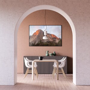 May include: A dining room with a white table and four white chairs. The table is set with a black teapot and a green vase with a single flower. A framed photograph of a mountain range hangs on the wall behind the table. The room has a light brown wood floor and a white archway.