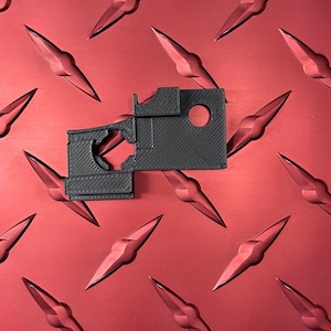 May include: A black plastic 3D printed part with a rectangular shape and a hole in the center. The part has a textured surface and appears to be a component for a larger assembly.