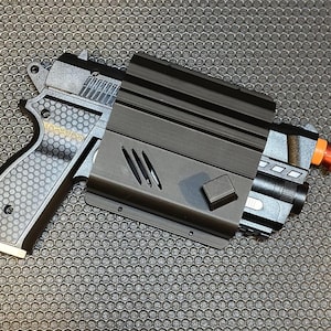 May include: A black and gray toy gun with a honeycomb-patterned grip and a black holster. The gun has a red and orange tip and a black cord attached to the bottom.