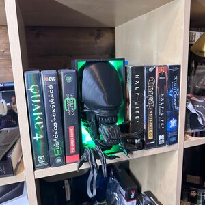 May include: A black and green 3D printed alien figure with glowing green eyes, used as a bookend on a wooden shelf. The alien is holding a book titled "Unreal Anthology" and is surrounded by other video game titles, including "Command & Conquer", "Quake 4", and "Half-Life 2".