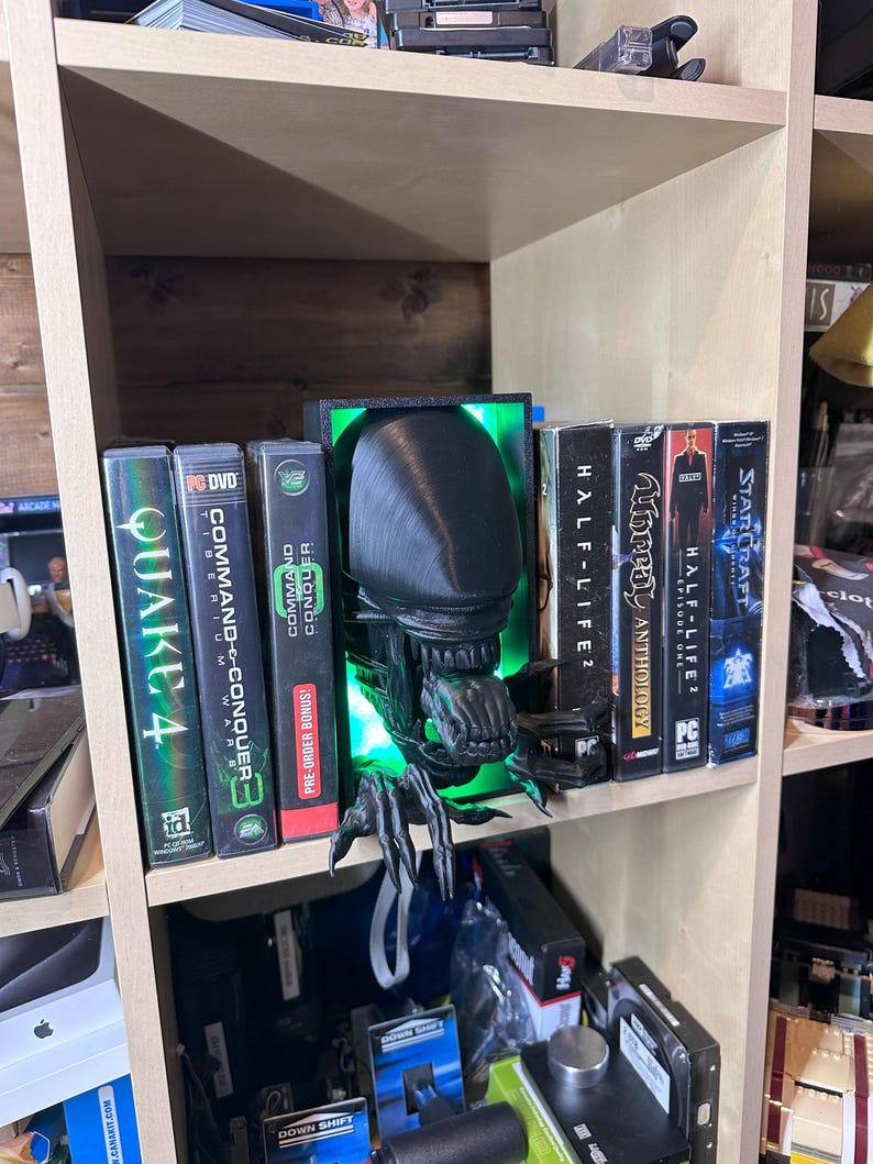 May include: A black and green 3D printed alien figure with glowing green eyes, used as a bookend on a wooden shelf. The alien is holding a book titled "Unreal Anthology" and is surrounded by other video game titles, including "Command & Conquer", "Quake 4", and "Half-Life 2".