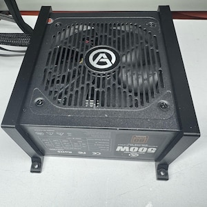 Op de afbeelding: Zwarte computer voeding met een ventilator en het merklogo in het midden. De voeding heeft een metalen rooster en de tekst "MOOG 500W" is zichtbaar. De unit heeft een zwart frame en montagebeugels.
