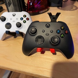 May include: Two Xbox Series X controllers, one white and one black, are displayed on a wooden surface. The black controller has red buttons and a stand.