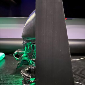 May include: A black 3D printed figurine of an alien creature with green glowing eyes. The figurine is mounted on a black wooden base.
