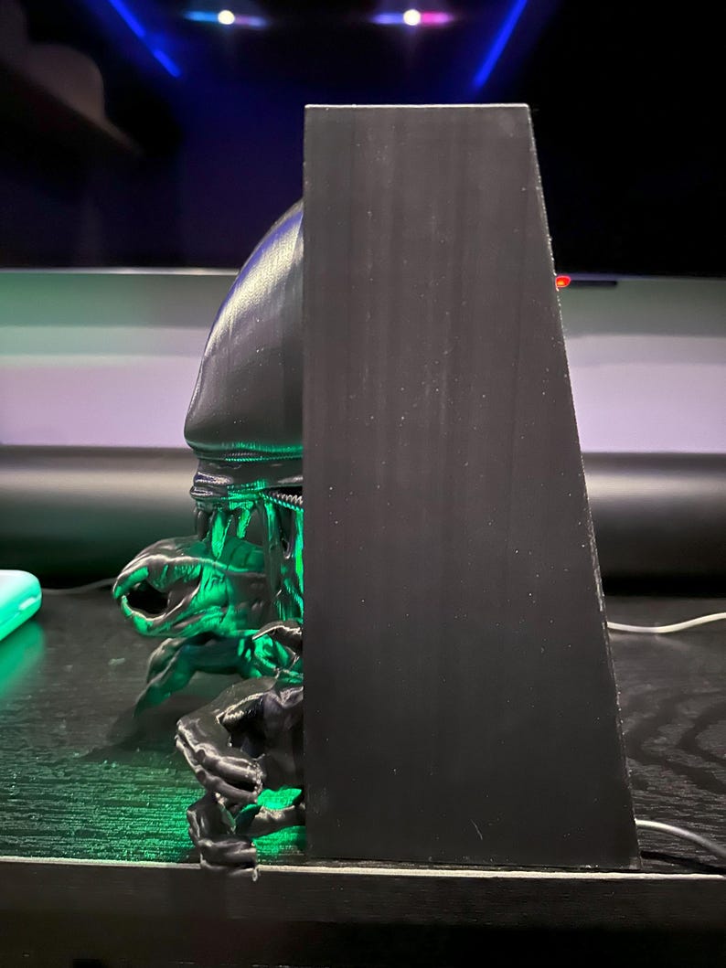 May include: A black 3D printed figurine of an alien creature with green glowing eyes. The figurine is mounted on a black wooden base.