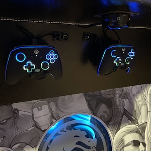May include: Two black Xbox controllers with blue and green lights, mounted on a wall with a Mortal Kombat logo behind them.