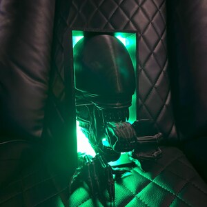 May include: A 3D printed alien bust with a green light shining through the head and mouth. The bust is in a black frame and is sitting on a black leather surface.