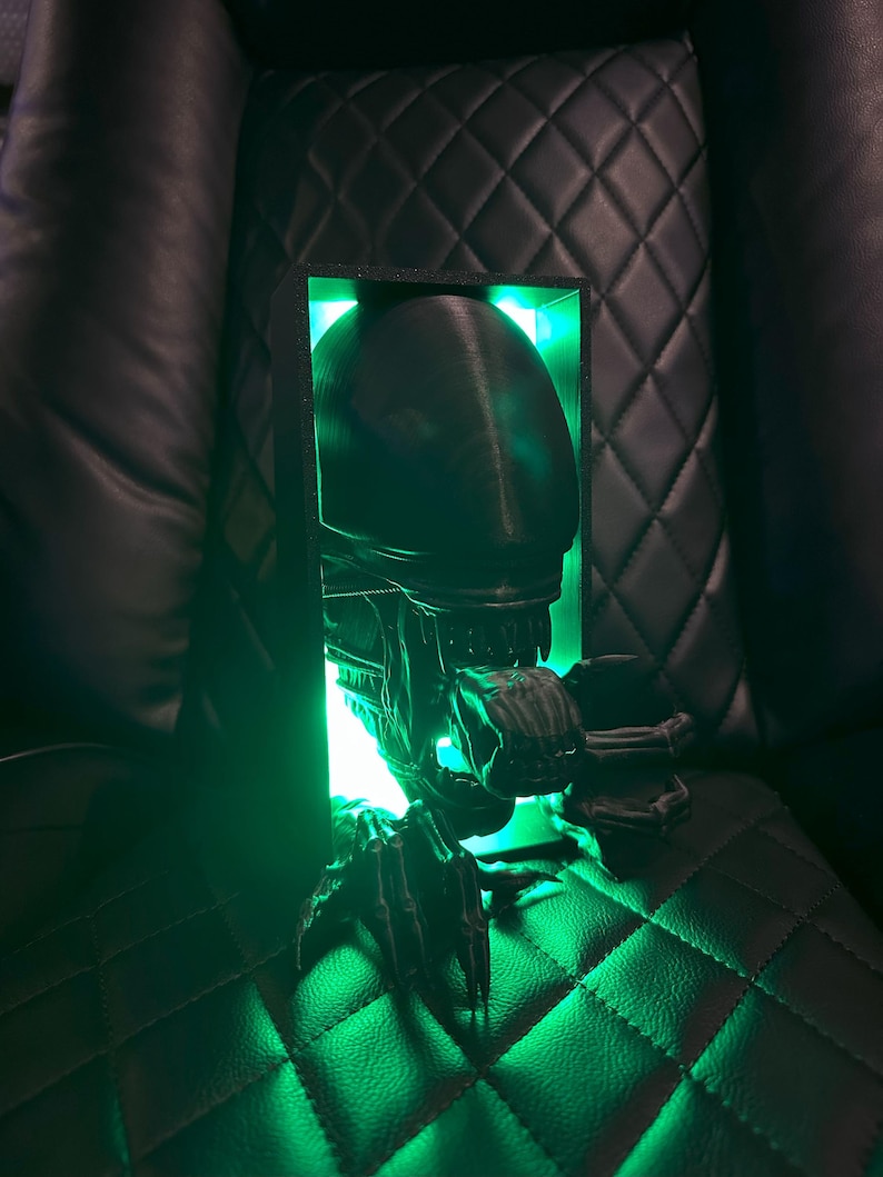 May include: A 3D printed alien bust with a green light shining through the head and mouth. The bust is in a black frame and is sitting on a black leather surface.