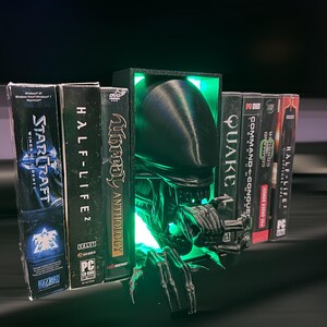 May include: A black and green 3D printed bookend shaped like an alien creature with glowing green eyes. The bookend is holding up a row of video game cases, including StarCraft, Half-Life 2, Unreal Anthology, Quake, and Command & Conquer.