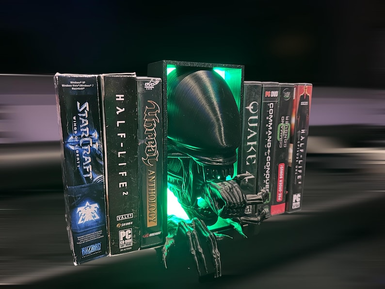 May include: A black and green 3D printed bookend shaped like an alien creature with glowing green eyes. The bookend is holding up a row of video game cases, including StarCraft, Half-Life 2, Unreal Anthology, Quake, and Command & Conquer.