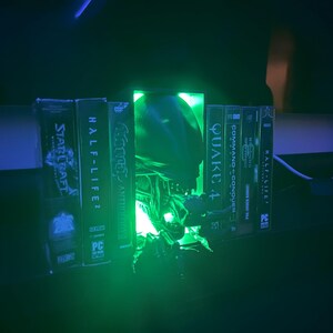 May include: A green glowing light illuminates a collection of video game cases, including StarCraft, Half-Life 2, Quake 4, Command & Conquer, and Half-Life. The light is coming from a green alien head sculpture.