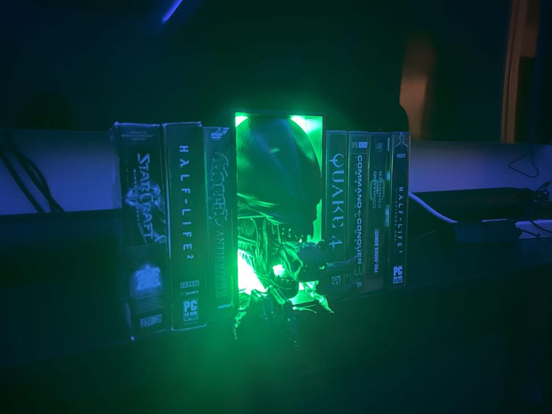May include: A green glowing light illuminates a collection of video game cases, including StarCraft, Half-Life 2, Quake 4, Command & Conquer, and Half-Life. The light is coming from a green alien head sculpture.