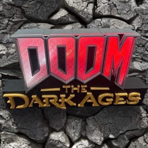 May include: A 3D sign featuring the text "DOOM THE DARK AGES". The word "DOOM" is in large, red, block letters with a silver outline. The text "THE DARK AGES" is in gold, stylized lettering. The sign is set against a stone background.