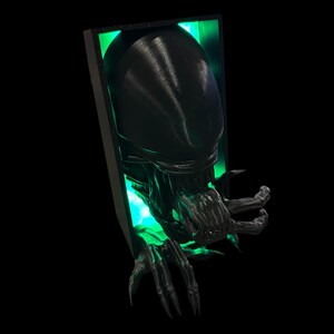 May include: A black 3D printed alien bust with glowing green eyes, emerging from a black frame. The alien has a large head with a mouth full of sharp teeth and long, clawed fingers.