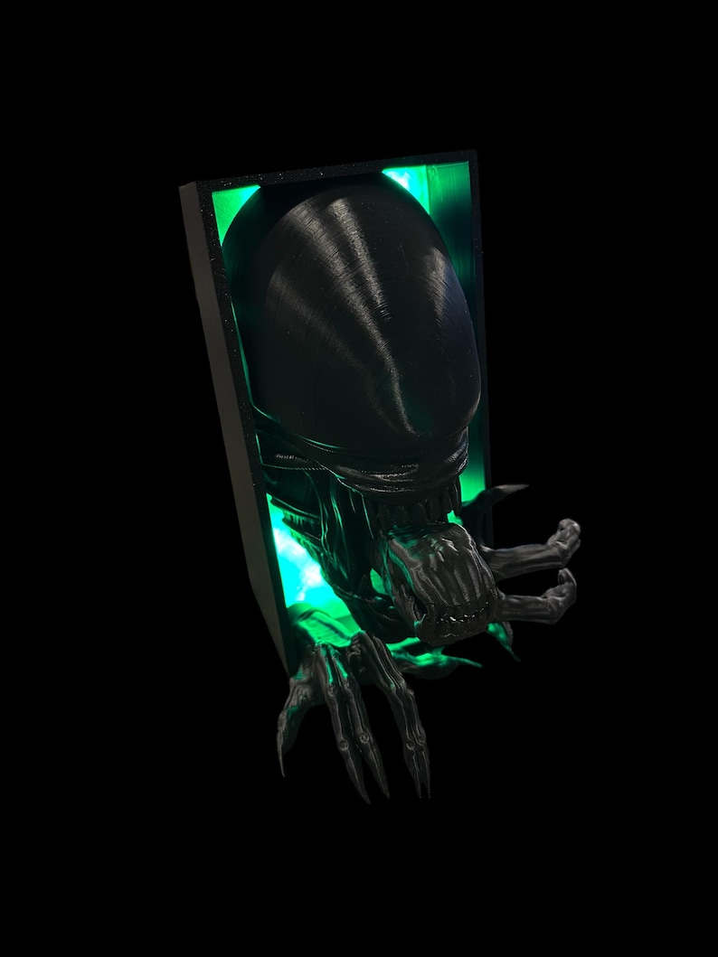 May include: A black 3D printed alien bust with glowing green eyes, emerging from a black frame. The alien has a large head with a mouth full of sharp teeth and long, clawed fingers.