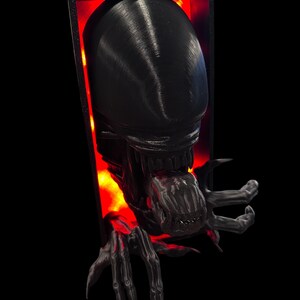 May include: A black 3D printed alien bust with a glowing red background. The alien has a large head with a gaping mouth and sharp teeth. The alien's hands are reaching out from the frame.