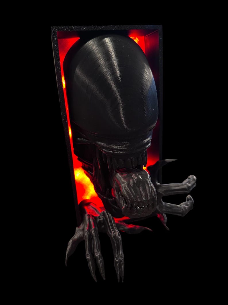 May include: A black 3D printed alien bust with a glowing red background. The alien has a large head with a gaping mouth and sharp teeth. The alien's hands are reaching out from the frame.