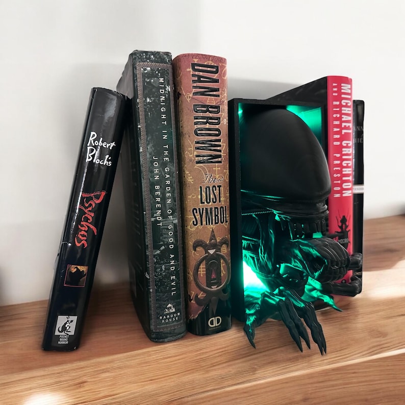 May include: Black bookends shaped like an alien creature with glowing green eyes. The bookends are holding up a book titled "The Lost Symbol" by Dan Brown, a book titled "Midnight in the Garden of Good and Evil" by John Berendt, and a book titled "Psychos" by Robert Bloch.