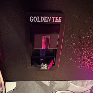 May include: A black plastic slot with the words "GOLDEN TEE" printed in white. The slot has a red light shining through it.