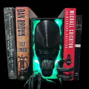 May include: Black and green bookends shaped like an alien creature with glowing green eyes. The bookends are holding up two books, one titled "The Lost Symbol" by Dan Brown and the other titled "Micro" by Michael Crichton.
