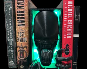 May include: Black and green bookends shaped like an alien creature with glowing green eyes. The bookends are holding up two books, one titled "The Lost Symbol" by Dan Brown and the other titled "Micro" by Michael Crichton.