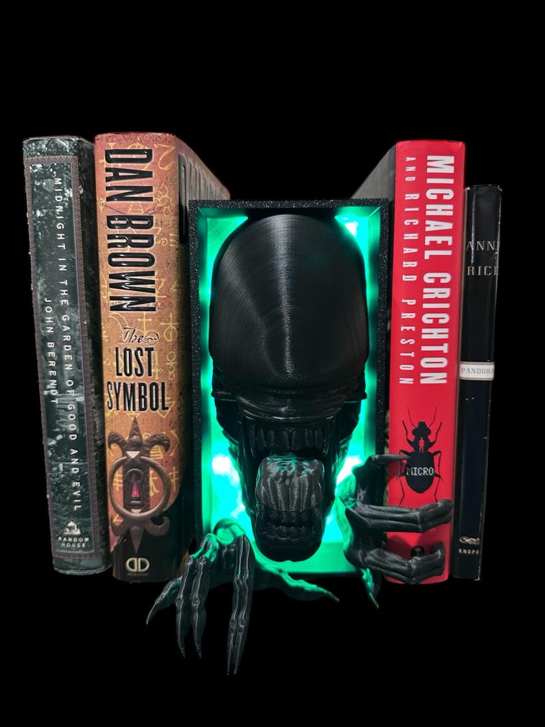 May include: Black and green bookends shaped like an alien creature with glowing green eyes. The bookends are holding up two books, one titled "The Lost Symbol" by Dan Brown and the other titled "Micro" by Michael Crichton.