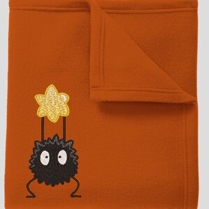 May include: A plush orange blanket with an embroidered black and white Totoro holding a yellow star.