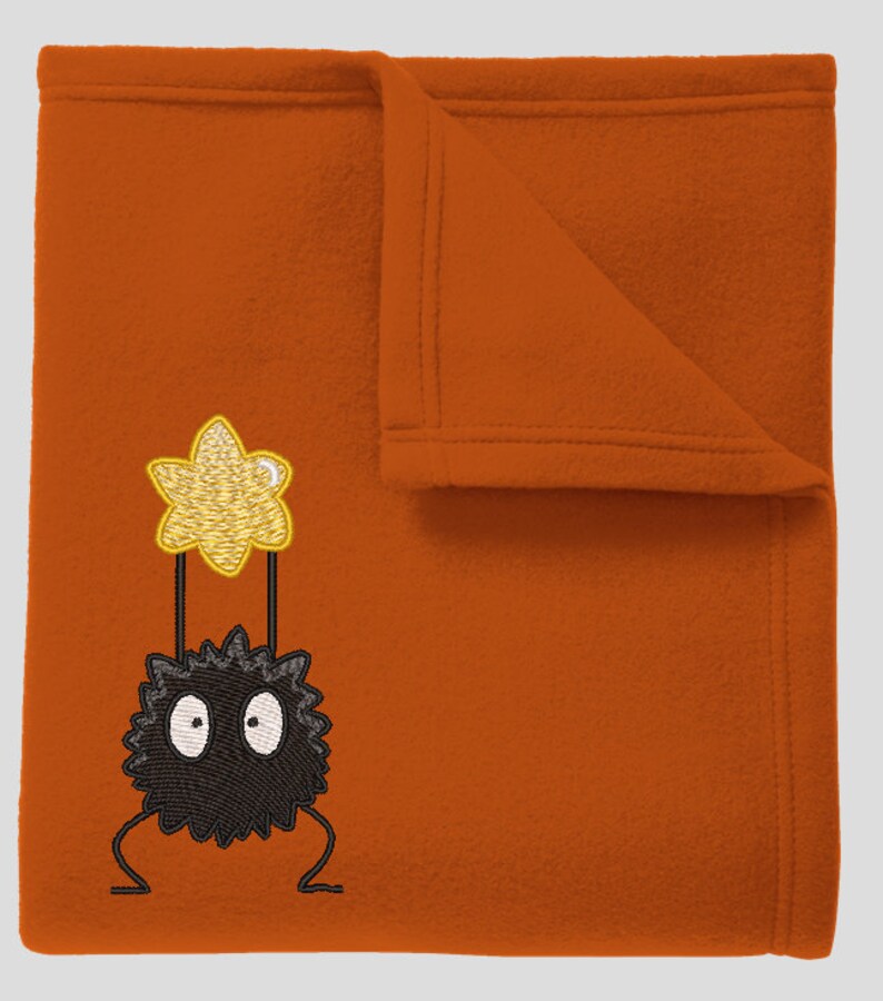 May include: A plush orange blanket with an embroidered black and white Totoro holding a yellow star.