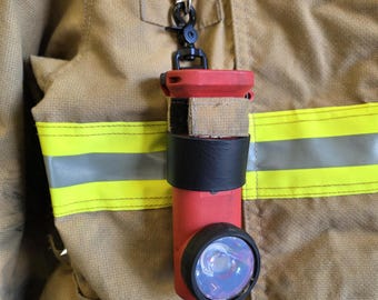 May include: A red and black flashlight attached to a tan firefighter's coat with yellow reflective stripes. The flashlight is cylindrical with a black bezel around the lens and a silver clip.
